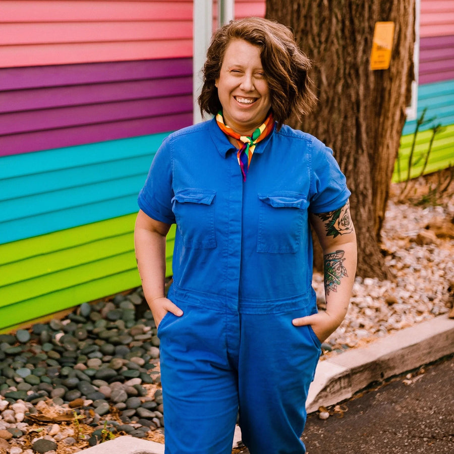 A smiling woman stands outdoors wearing a bright blue short-sleeve jumpsuit with a colorful neck scarf. She is posed in front of a colorful striped wall and a tree trunk.