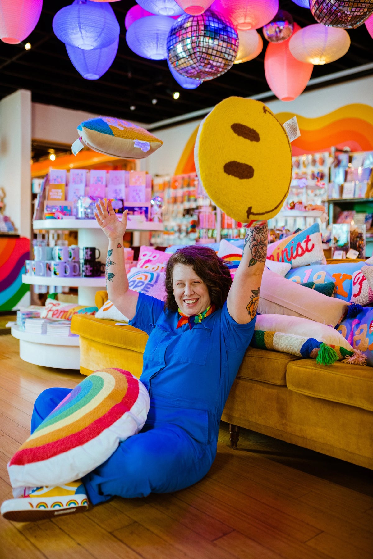 A woman in a blue outfit sits on the floor of a colorful retail store, holding a large yellow smiley face pillow and tossing a rainbow-striped pillow in the air. She is surrounded by plush pillows, a mustard-yellow sofa, and hanging paper lanterns and disco balls on the ceiling.