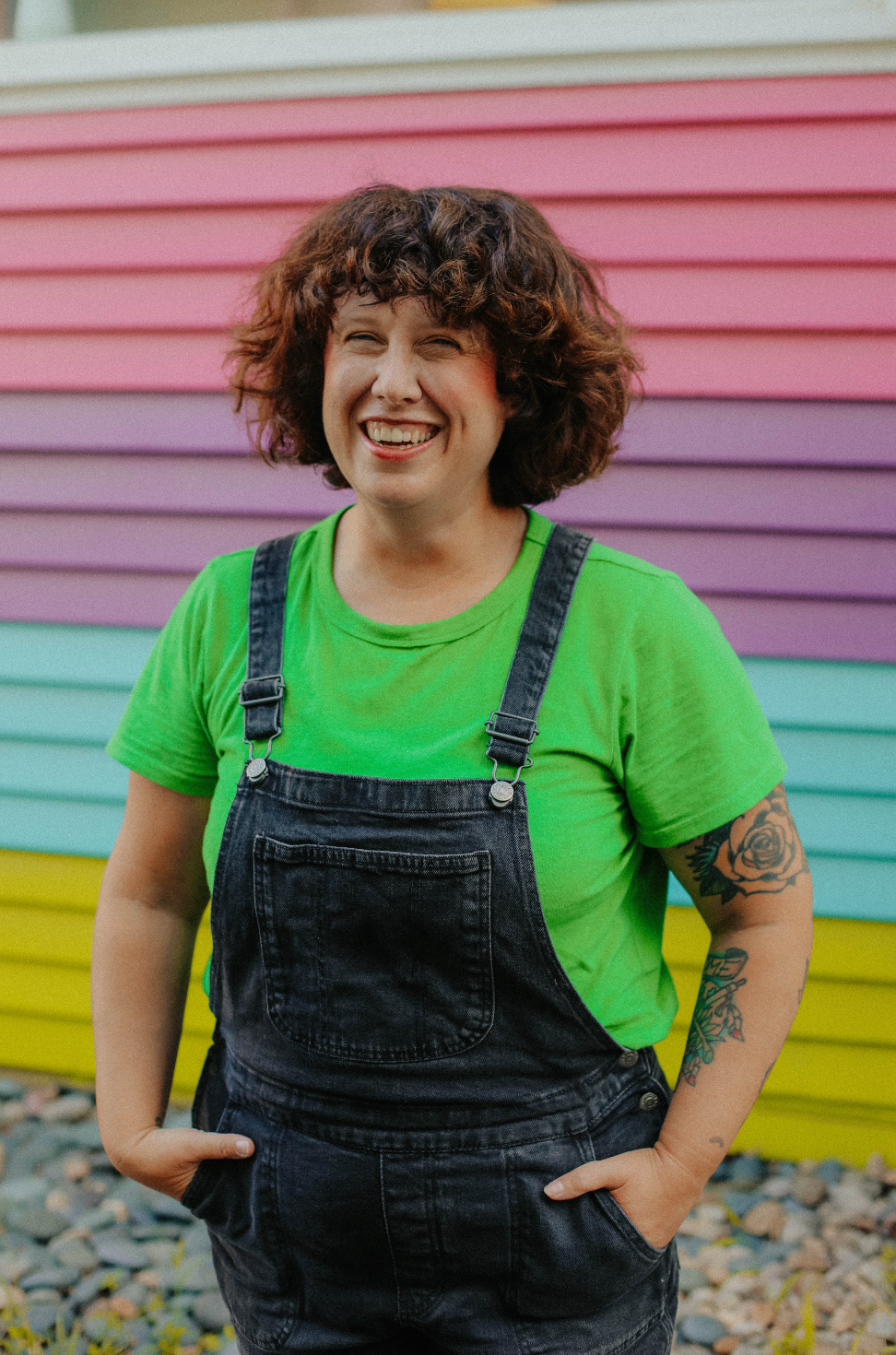 A smiling person with curly brown hair stands in front of a colorful striped wall, wearing a bright green T-shirt and dark denim overalls with hands in the pockets.